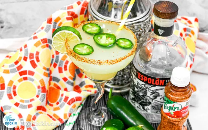 Spicy Margarita with jalapeno slices and lime wedge fo garnish. Cocktail shaker, bottle of Tequila and Tajin chili lime seasoning in background.