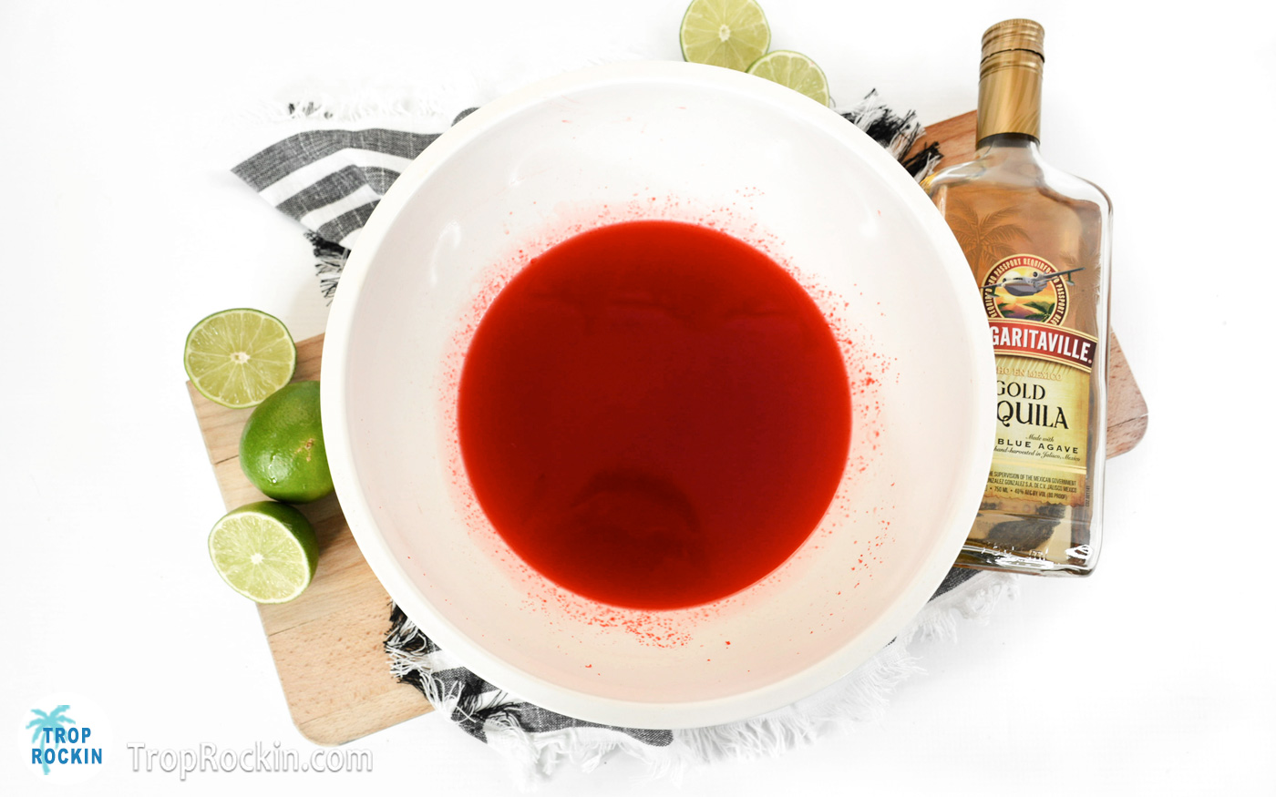 Strawberry jello mixed with water and tequila in a large bowl with a bottle of tequila and fresh limes in background.