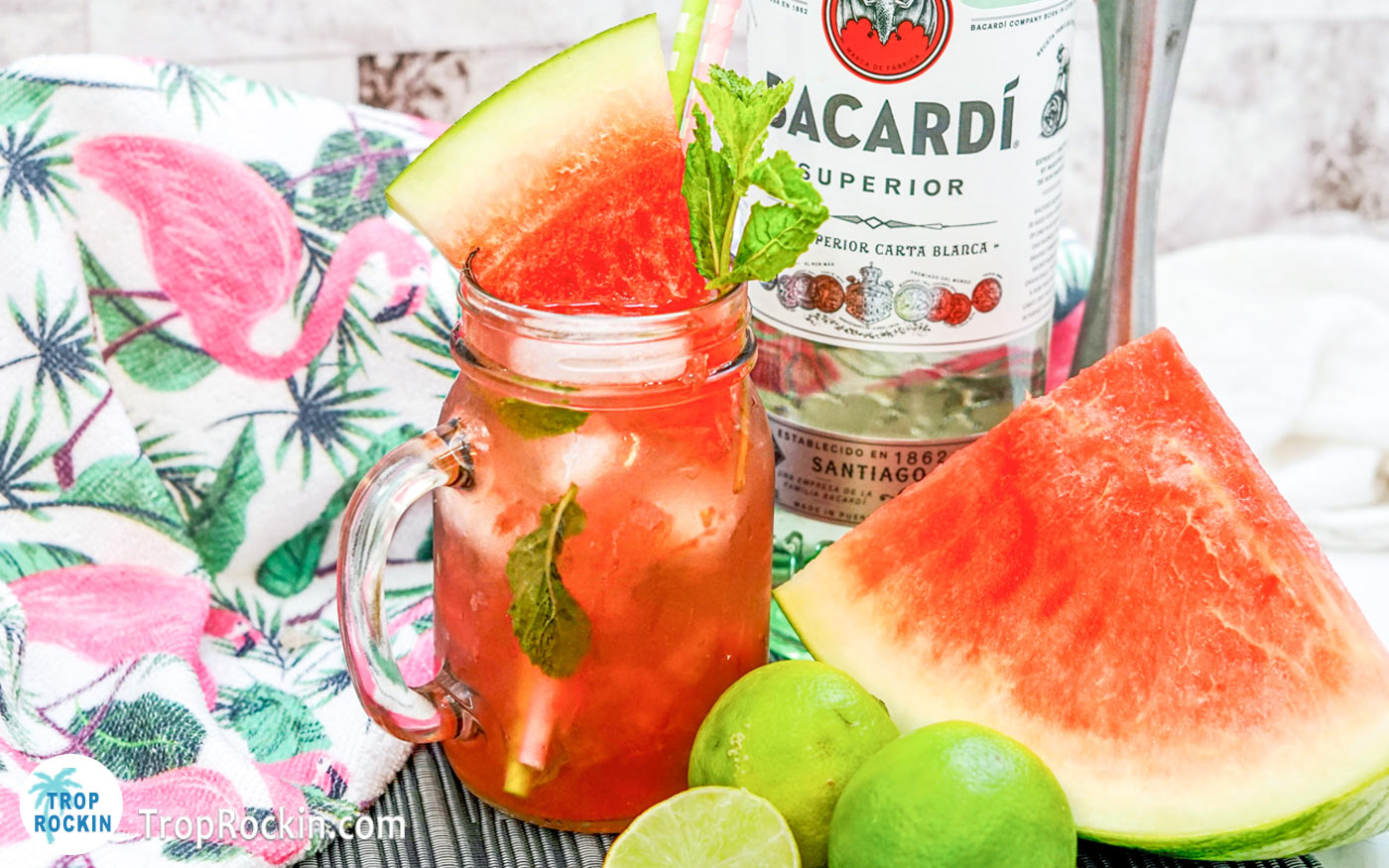 Watermelon Mojito with a sprig of mint and watermelon wedge for garnish in a mason jar. A bottle of Bacardi Rum in the background with several limes and a large watermelon slice.