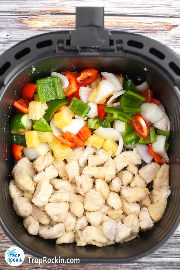 Air fryer basket filled with chicken on one side and bell pepper, onion and pineapple on the other half.