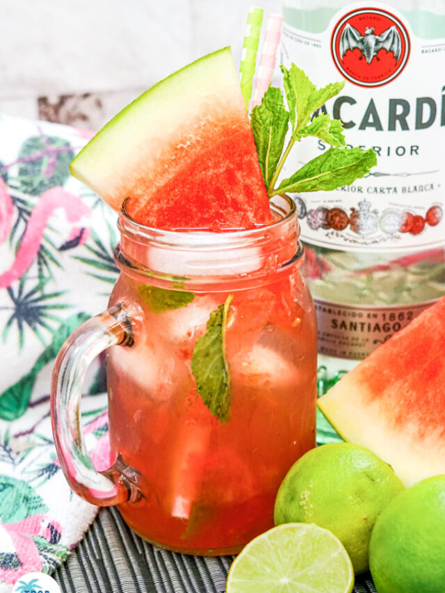 Watermelon Mojito with a sprig of mint and watermelon wedge for garnish in a mason jar. A bottle of Bacardi Rum in the background with several limes and a large watermelon slice.