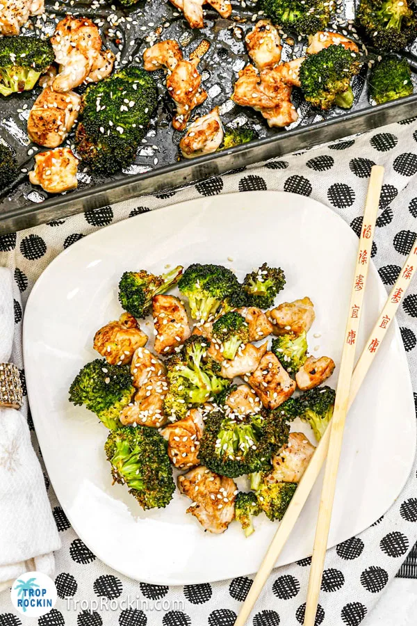Air Fryer Broccoli and Chicken on a white plate with a white napkin rolled up with a black and white polka dot kitchen towel underneath. Partial view of chicken and broccoli in the air fryer basket in the background.