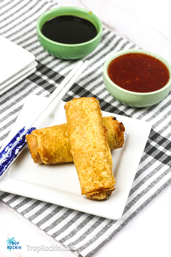 Two air fried egg rolls on small white plate with dipping sauce in the background.