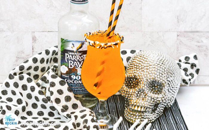 Orange Halloween Punch Cocktail with Halloween sprinkles on the rim of the glass and two orange and black straws. Coconut rum bottle and a sparkly skull in the background.