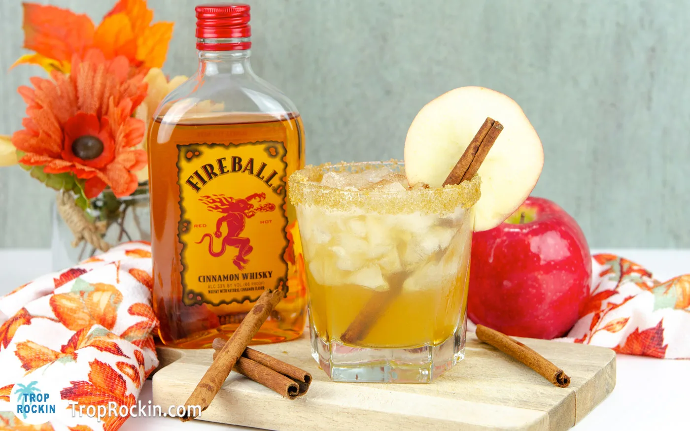 Apple pie cocktail in a lowball glass with a brown sugar rim, cinnamon stick and apple wheel for garnish sitting on a cutting board with Fireball Cinnamon Whisky and a red apple in the background.