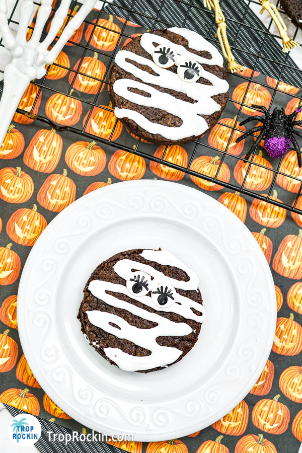 Halloween mummy brownie on a white plate with another mummy decorated brownie on a wire rack / cooling rack.