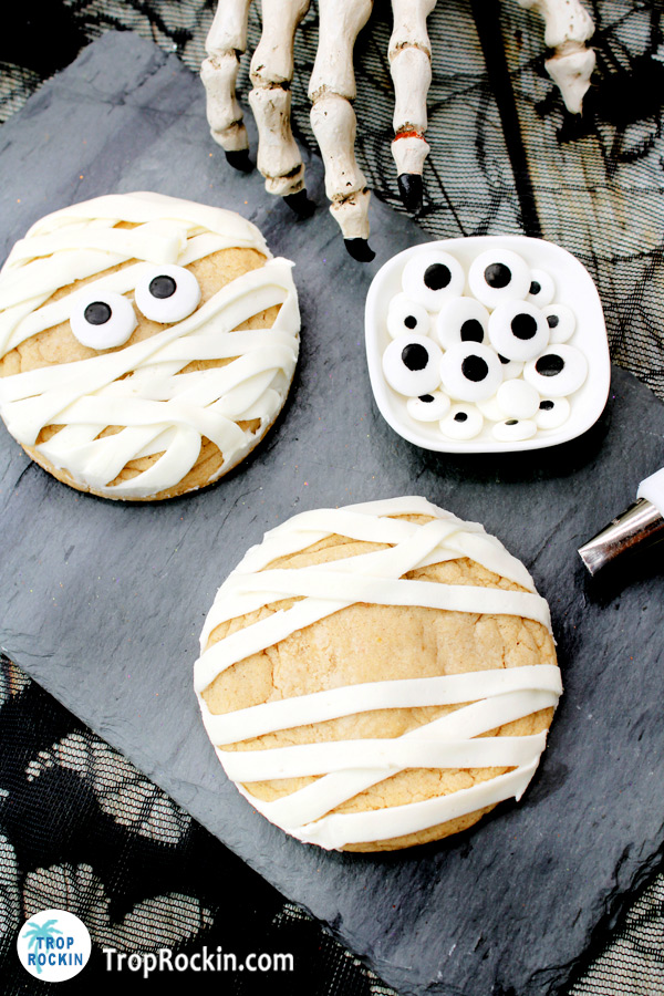 A maple cinnamon cookie with mummy stripes of frosting in foreground with a mummy cookie and bowl of candy eyeballs in background.