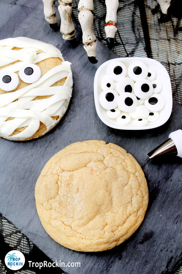 A maple cinnamon cookie without decorations in foreground with a mummy cookie and bowl of candy eyeballs in background.