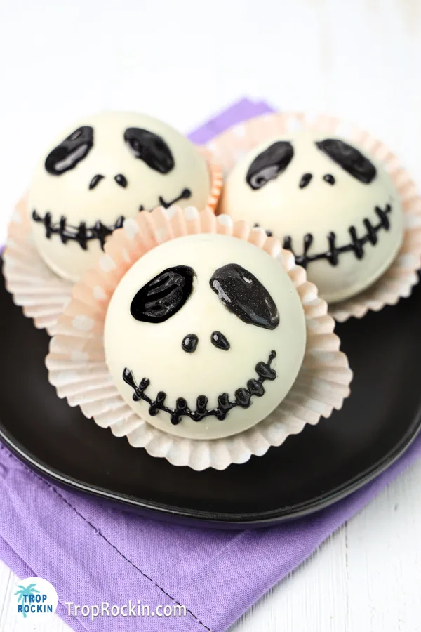 Three Jack Skellington hot chocolate bombs on a black plate with a purple napkin under the plate.