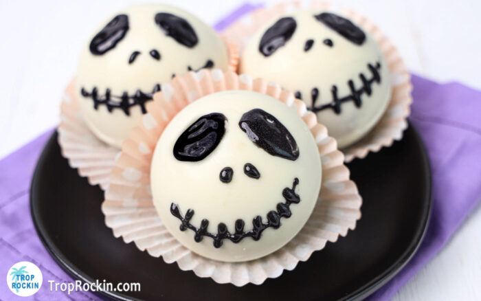 Three Jack Skellington hot chocolate bombs on a black plate with a purple napkin under the plate.