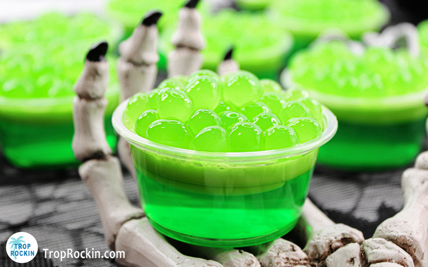 A witches brew jello shot propped on a skeleton hand with witches brew jello shots in the background.
