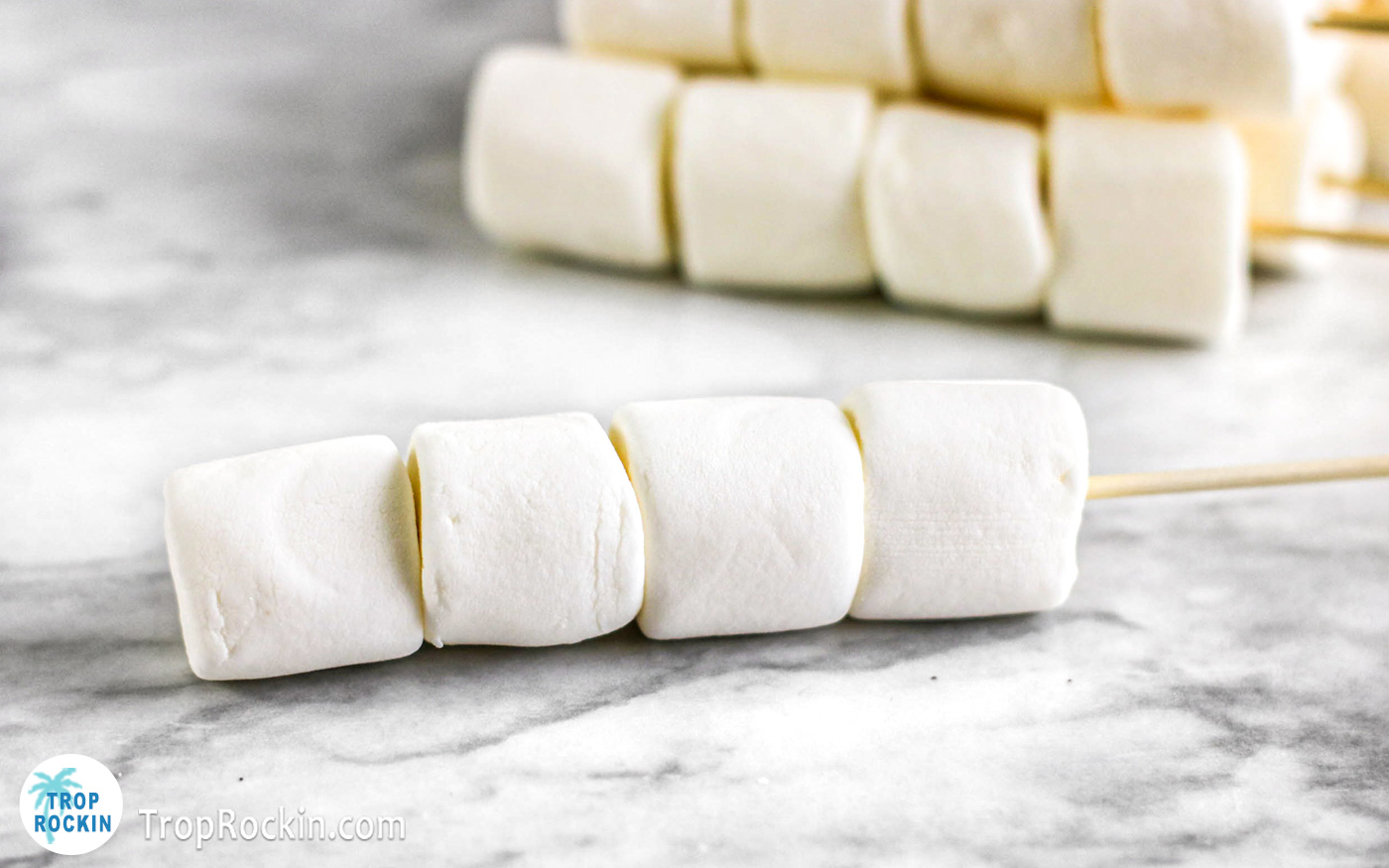 Four jumbo marshmallows on a stick laying on the counter top.