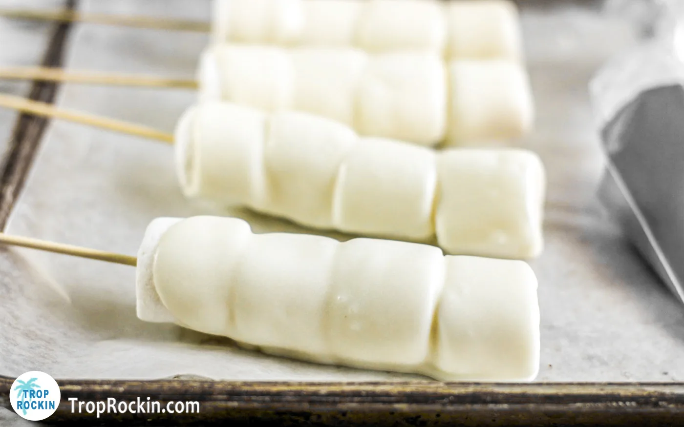 Marshmallow pops covered in melted white chocolate on a baking sheet covered with parchment paper.