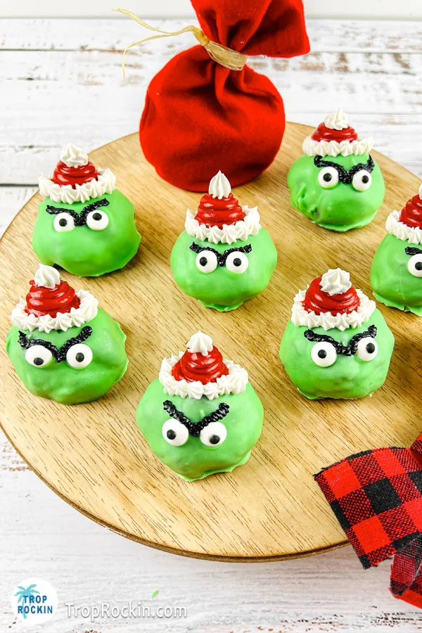 Grinch Oreo Balls with red santa hats, candy eyeballs and dark eyebrows displayed on a round cutting board.