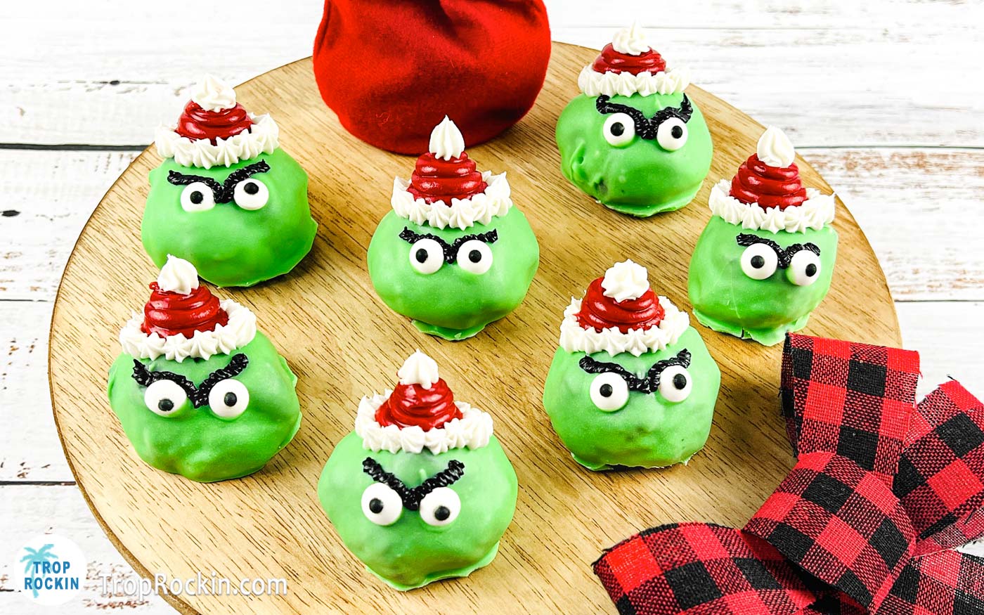 Grinch Oreo Balls with red santa hats, candy eyeballs and dark eyebrows displayed on a round cutting board.