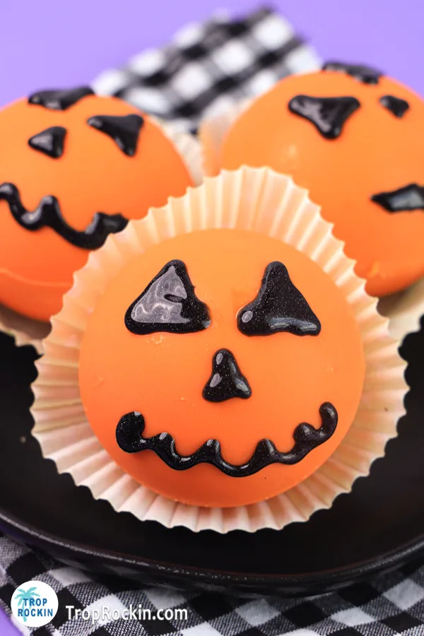 Three halloween hot chocolate bombs decorated with a hack-o-lantern face in cupcake liners on a plate.