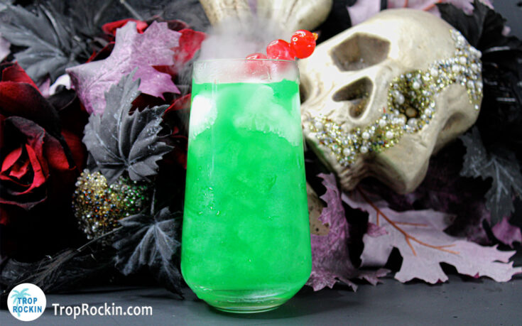 Witches Brew Cocktail, bright green drink in a tall glass with maraschino cherries for garnish with halloween decor in the background.
