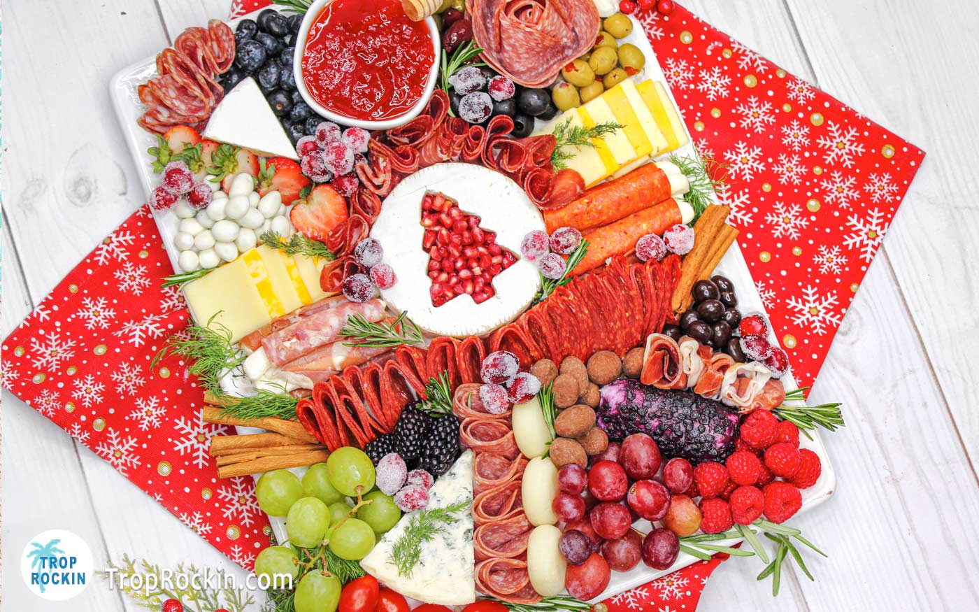 Christmas Charcuterie Board with a large variety of meats, cheeses, fresh fruits and snacks dispayed on a white platter with a red kitchen towel underneath on a white wood board background.