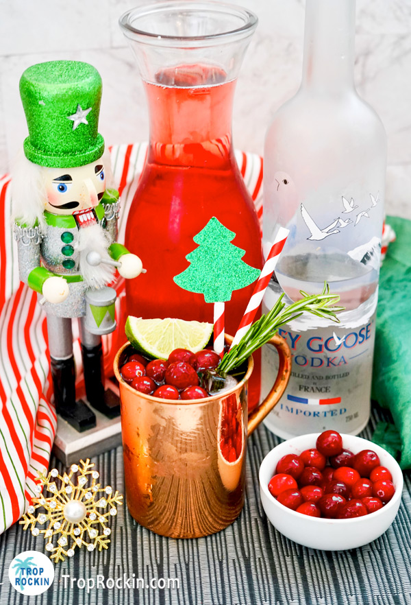 Christmas mule drink with a caraft of cranberry juice and bottle of vodka in the background along with a small bowl of fresh cranberries and festive christmas decorations.