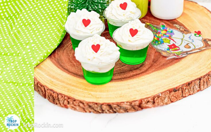 Grinch Jello Shots with whipped cream topping and red heart sprinkles sitting on a wood cutting board.