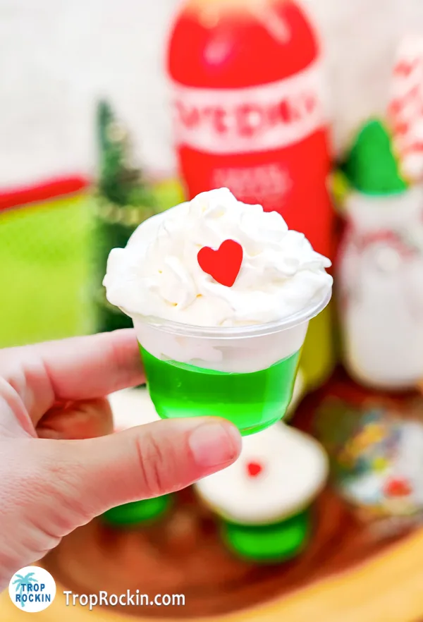 Holding a single Grinch Jello Shot with ingredients in the background.