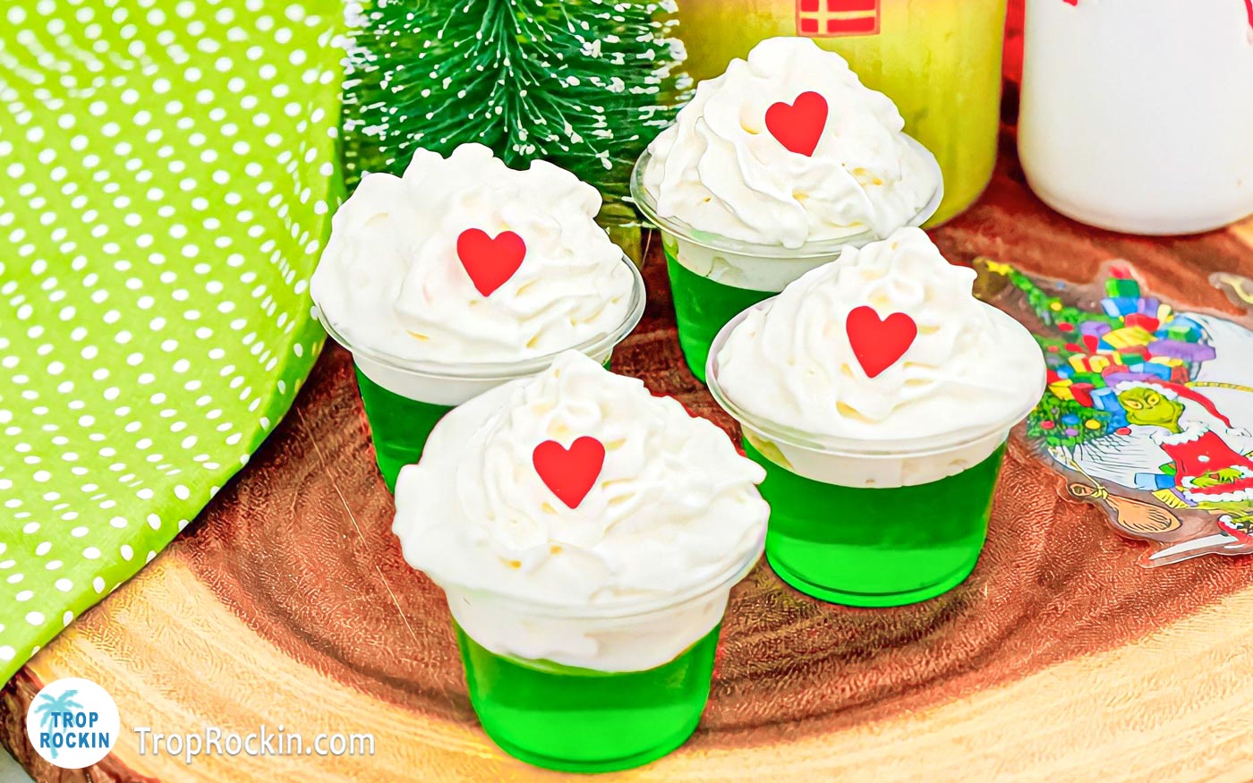 Four Grinch Jello Shots made with green jello and topped with whipped cream and a red heart sprinkle displayed on a wood cutting board.