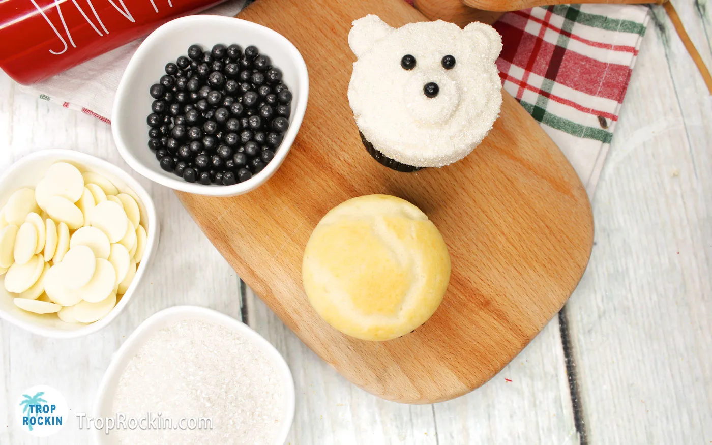 Basic Polar Bear Cupcake Ingredients displayed on wood background. Bowl of white chocolate wafers, sanding sugar, black pearl sprinkles, a vanilla cupcake and one fully decorated polar bear cupcake.