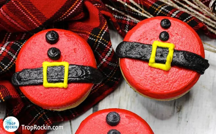 Two Santa Cupcakes with christmas plaid background decorated with frosting to look like Santa's belly with black frosting belt and buttons and a yellow frosting belt buckle and solid red frosting for the Santa Claus suit.