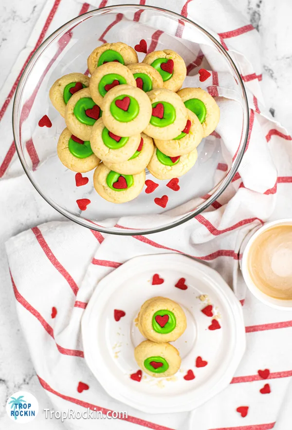 Top view of a Large clear bowl full of Grinch Thumbprint Cookies and a plate with a few cookies.