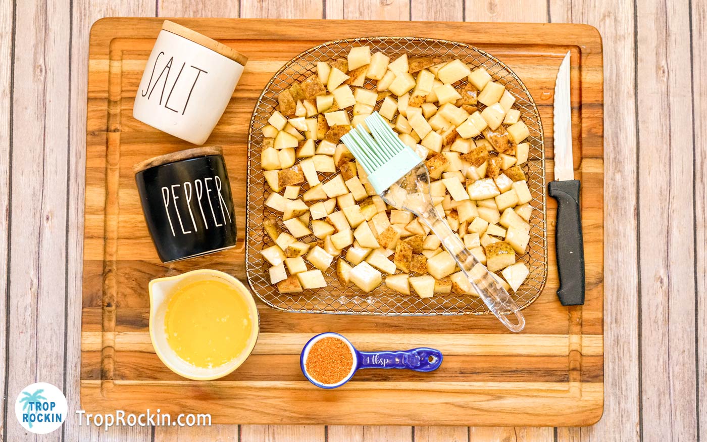 Cubed potatoes on air fryer tray with melted butter in a bowl to the side, using a basting brush to cover the potatoes with the butter.