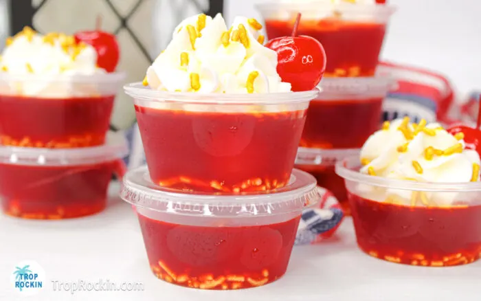 Cherry Jello Shots with whipped cream, a cherry and sprinkles displayed on counter top.
