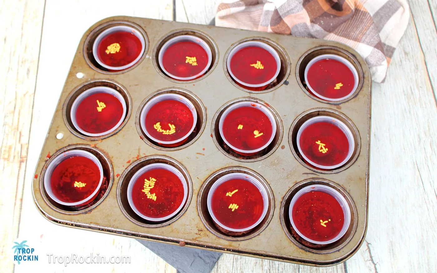 Filled jello shot cups with gold sprinkles sitting inside a muffin pan.