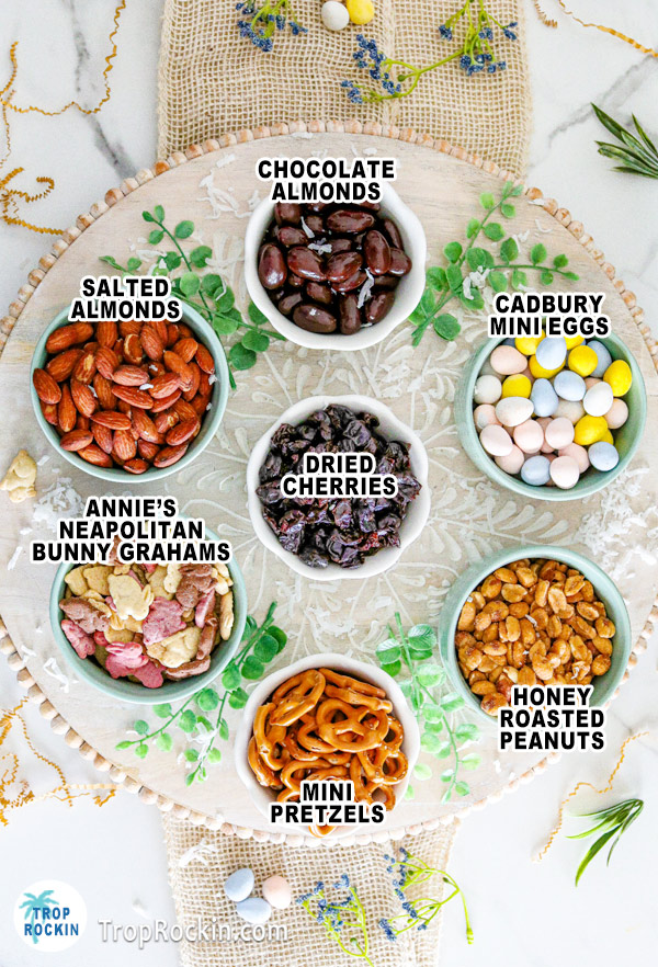 Easter Trail Mix ingredients in individual small bowls displayed on a platter and each ingredient is labeled with text overlay.