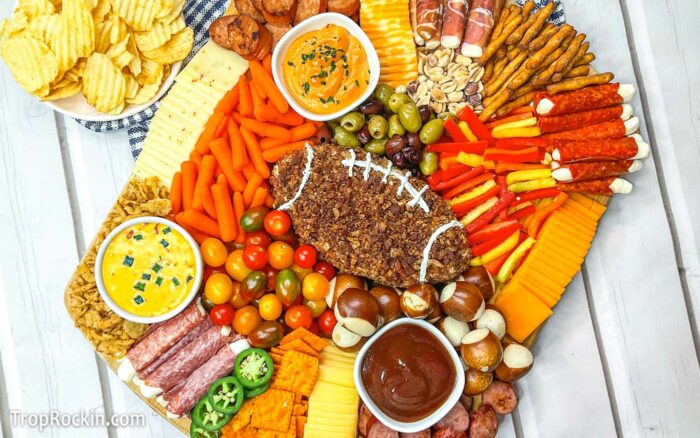 Football Charcuterie Board is a large rectangular cutting board filled with meats, cheeses, olives, vegetables, crackers and dips. The center cheese ball is shaped like a football and decorated with sour cream to look like football laces.