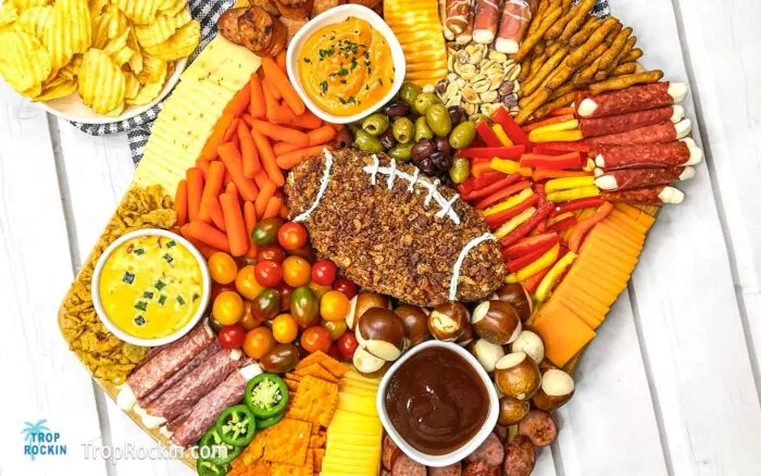 Football Charcuterie Board is a large rectangular cutting board filled with meats, cheeses, olives, vegetables, crackers and dips. The center cheese ball is shaped like a football and decorated with sour cream to look like football laces.