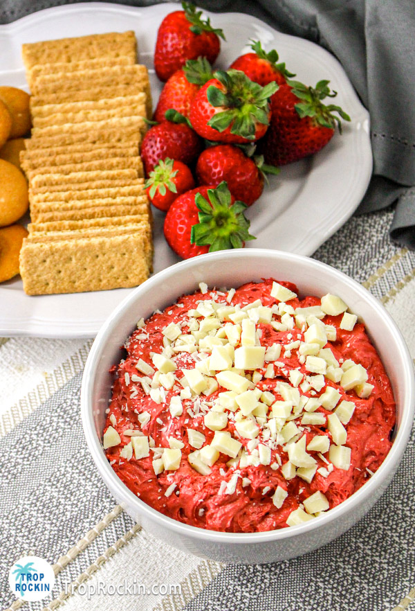White bowl of red velvet dip with white chocolate pieces on top. Plate of cookies and strawberries in the background.