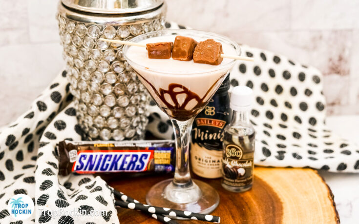 Snickers Drink in a martini glass with chocolate syrup and mini snickers bars for garnish with liquor and cocktail shaker in the background.