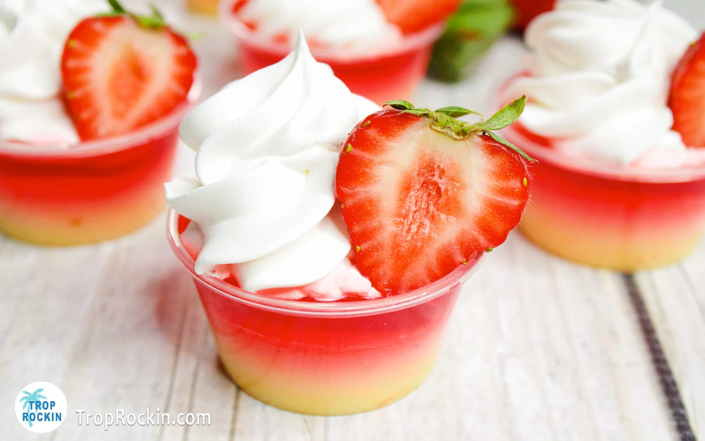 Strawberry Shortcake Jello Shots with two layers, bottom layer is vanilla pudding mixture, top layer strawberry jello mixture with whipped cream and a strawberry slice on top.