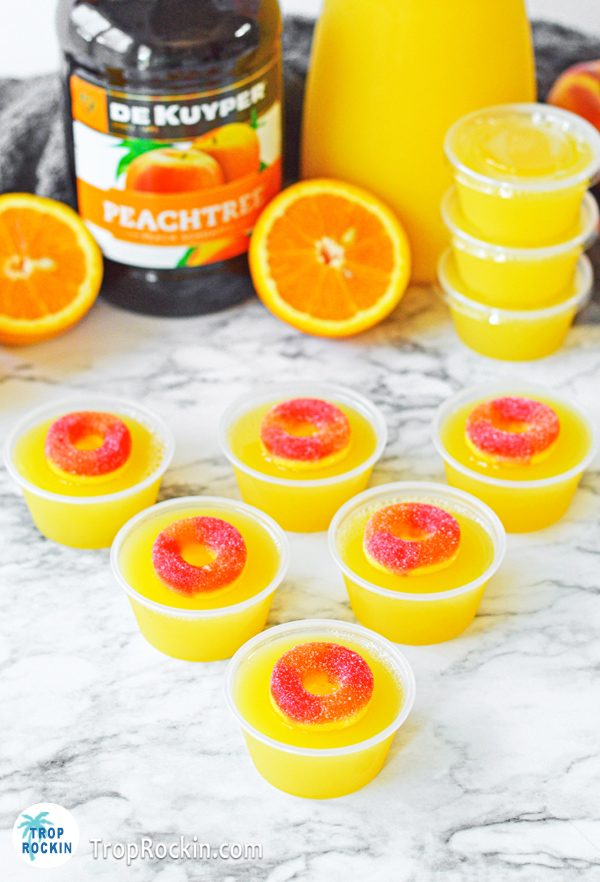 Fuzzy Navel Jello Shots with peach ring garnish displayed on marble counter top. Ingredient bottles in the background.