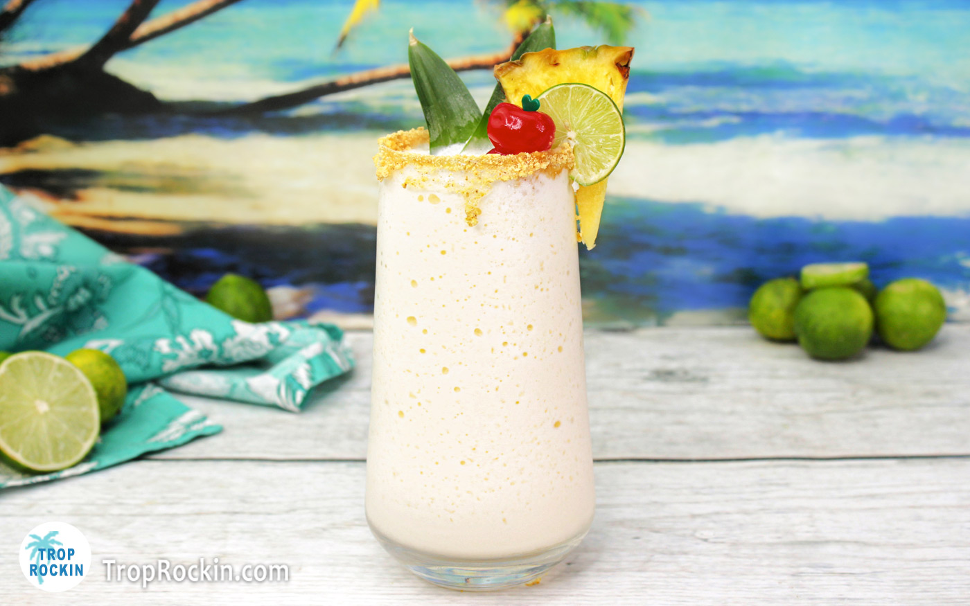 Key Lime Colada: Creamy white frozen drink with a pineapple slice, maraschino cherry, lime wheel and pineapple leaves for garnish. Graham cracker crumbs on the rim of the glass while sitting on a white wood counter top with a tropical beach background.
