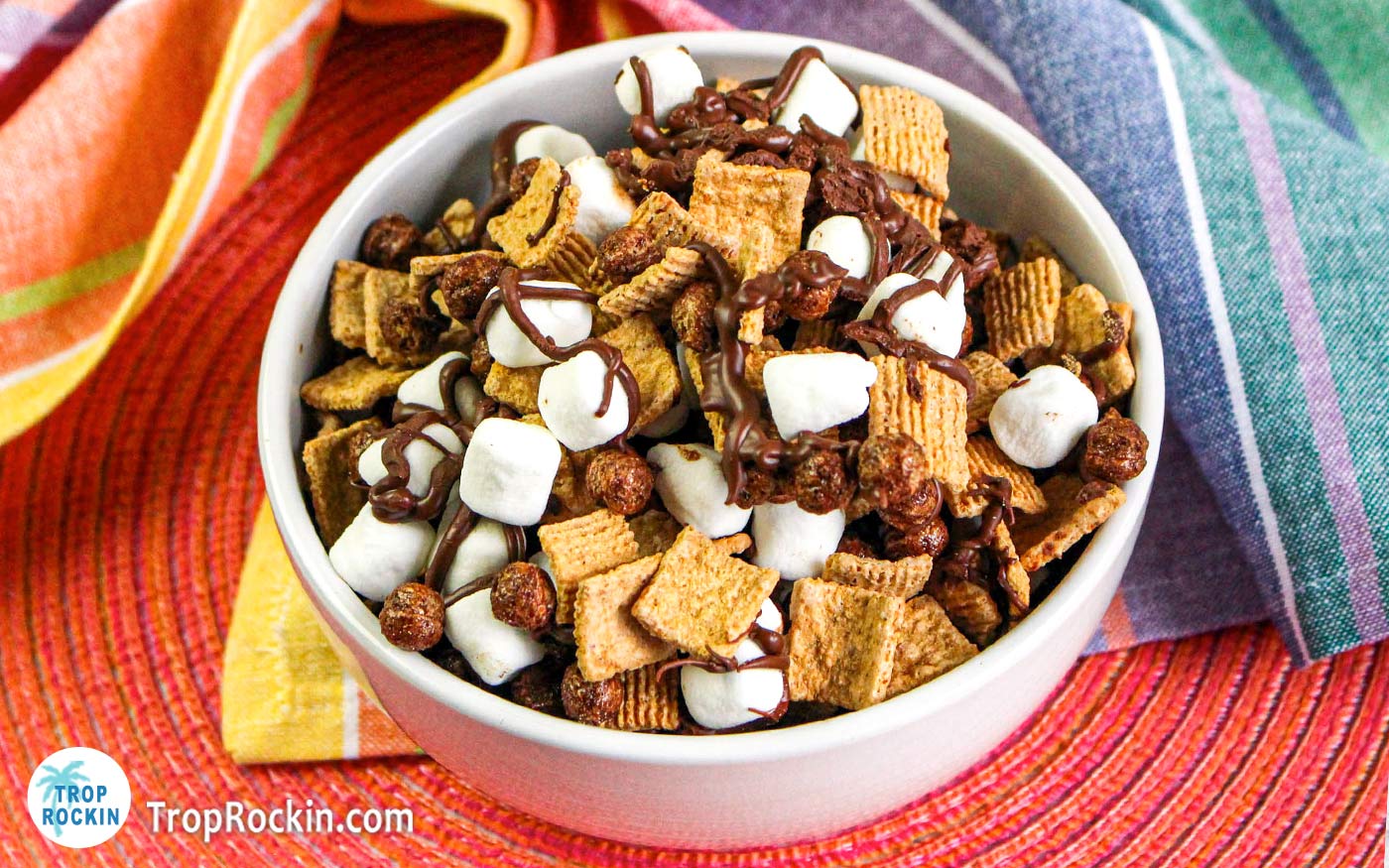 White bowl filled with s'mores snack mix on top of a bright pink place mat and a colorful kitchen towel.