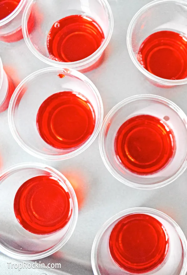 Jello shot cups lined up on a baking pan with cherry jello filling up 2/3 of the cup.