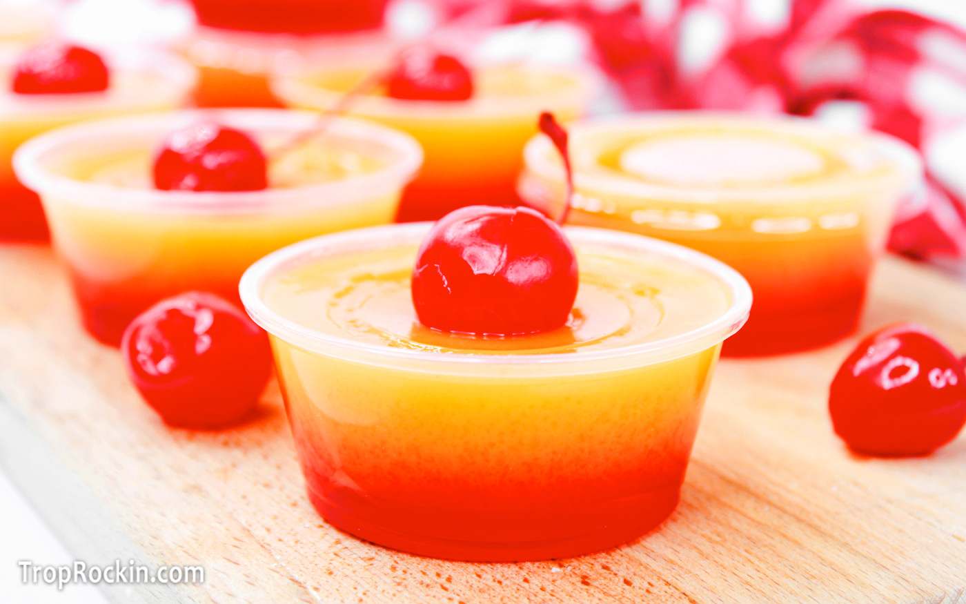 Tequila sunrise jello shots on a wood cutting board. Each jello shot has an orange layer on top and red layer on bottom with a maraschino cherry sitting on top.