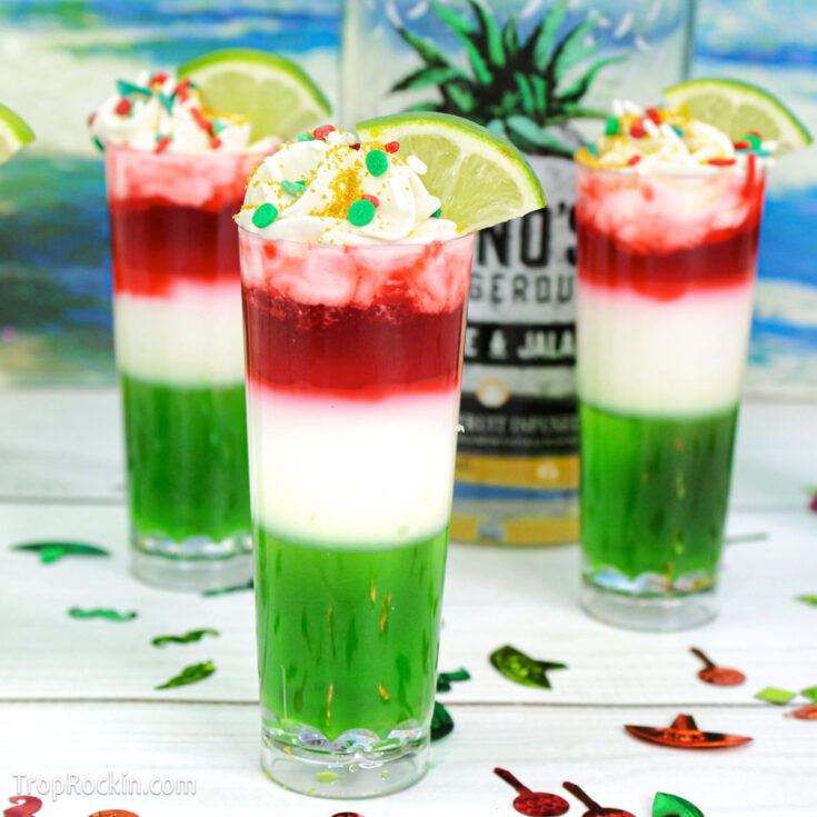 Three Mexican Flag Jello Shots with three layers: green, white and red. Topped with whipped cream and sprinkles and garnished with a lime wedge on a white wood tabletop with a tropical ocean background.