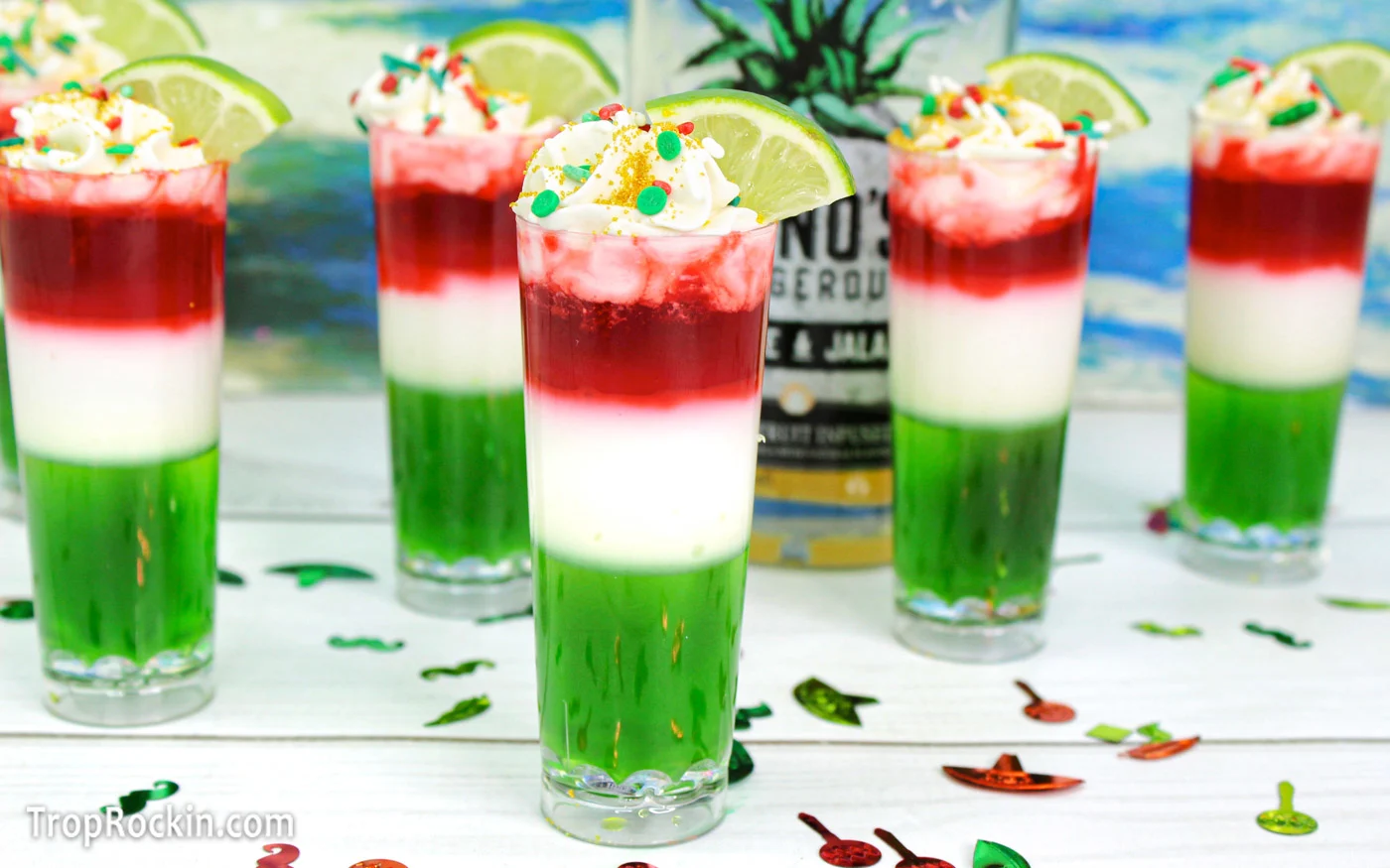 Five Mexican Flag Jello Shots with three layers: green, white and red. Topped with whipped cream and sprinkles and garnished with a lime wedge on a white wood tabletop with a tropical ocean background.