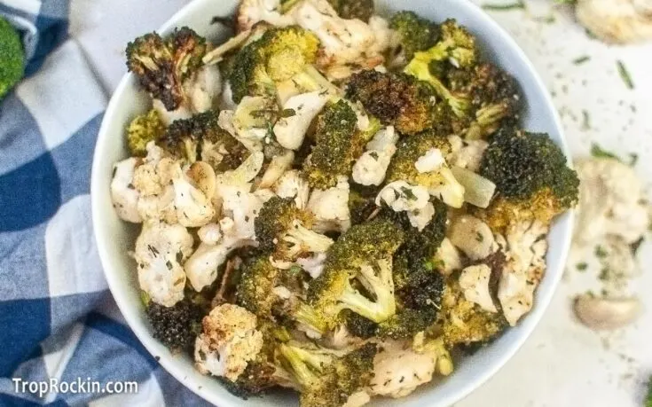 Bowl of air fryer broccoli and cauliflower seasoned and air fried on a white countertop with a blue and white kitchen towel.