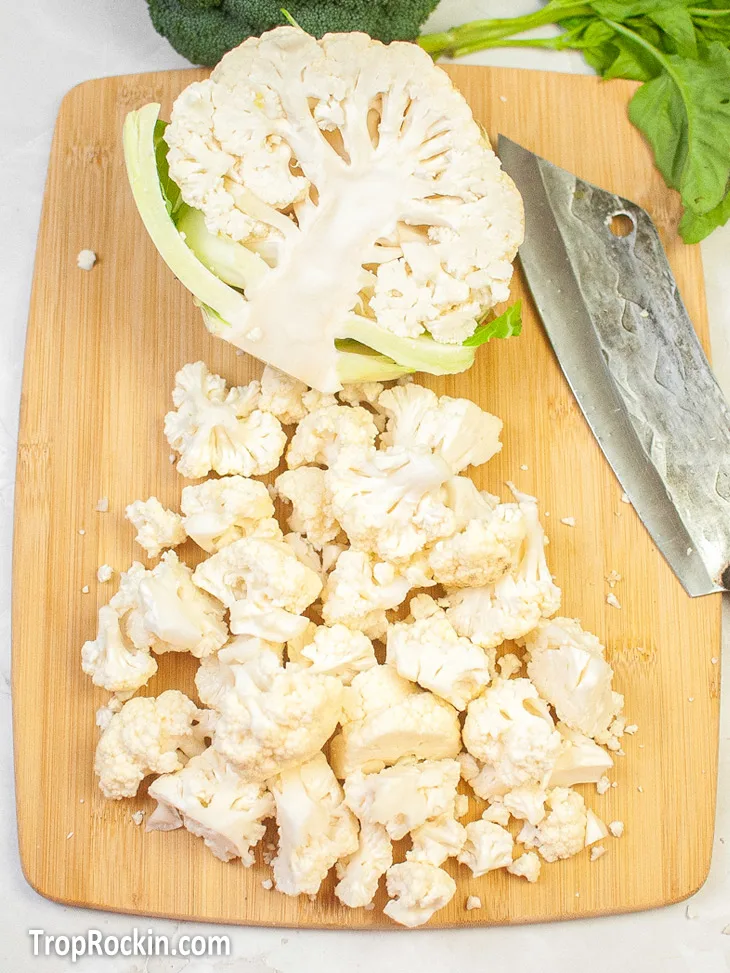 Cauliflower cut in half with half of it cut into cauliflower florets sitting on a wooden cutting board with a knife.