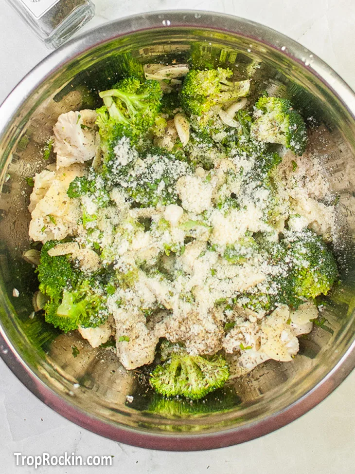 Stainless steel bowl with cut broccoli florets and cauliflower florets with oil, seasoning and Parmesan cheese poured on top.
