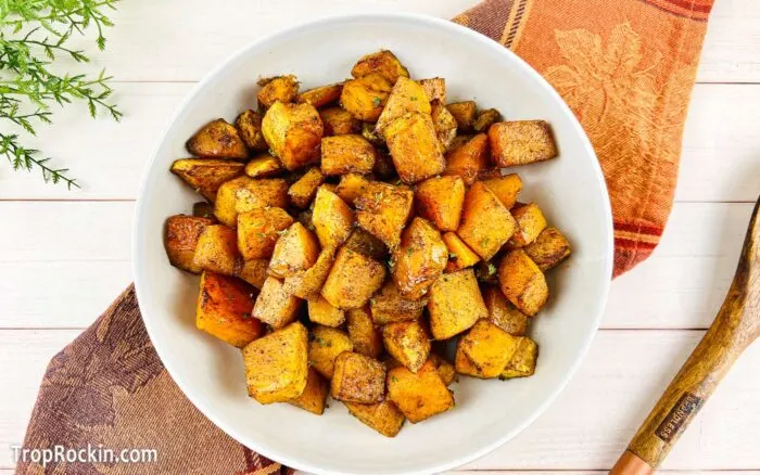 Cubed, seasoned and air fried, large white bowl of air fryer butternut squash on a white wood background with a fall colored linen napkin underneath the bowl.