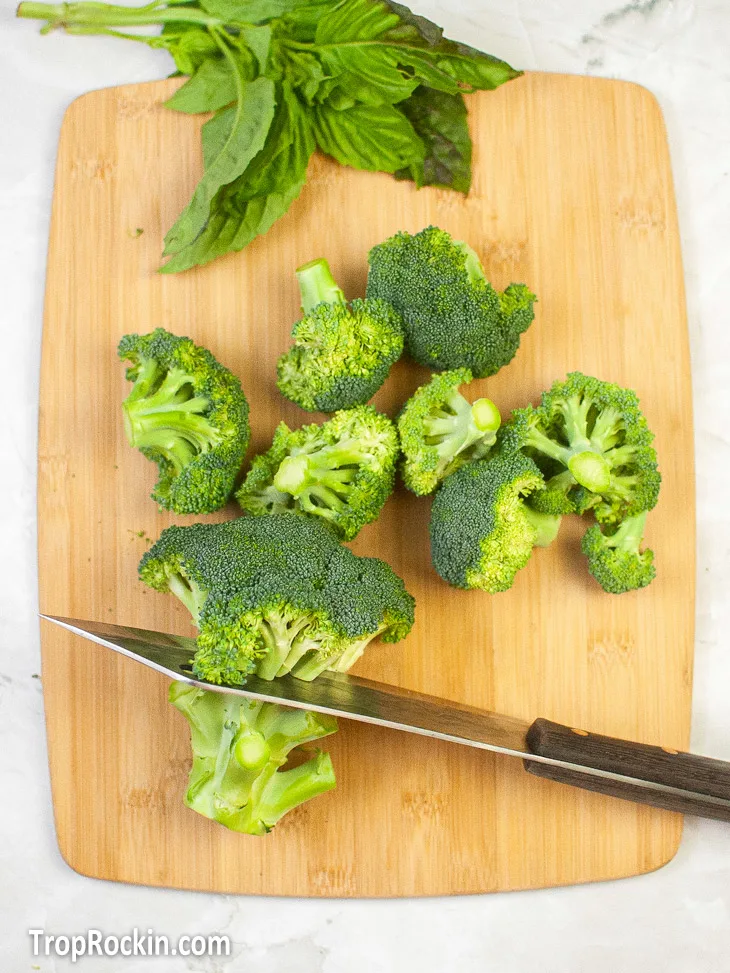 Cutting off stem of a broccoli floret with a knife while other already cut florets are also sitting on a wooden cutting board.